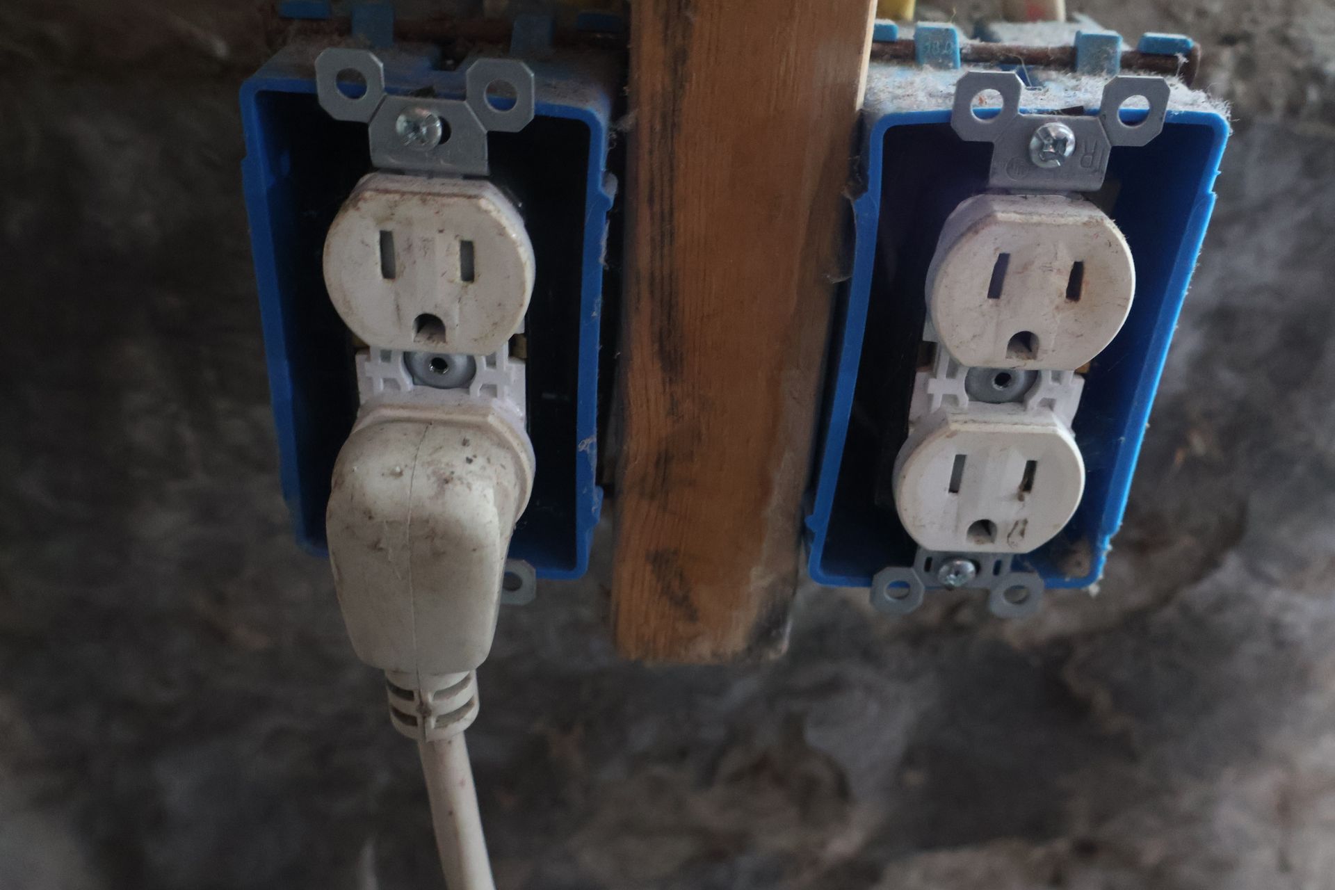 Two blue plastic electrical boxes mounted on a wooden stud, each containing a white duplex wall outlet with one plug.