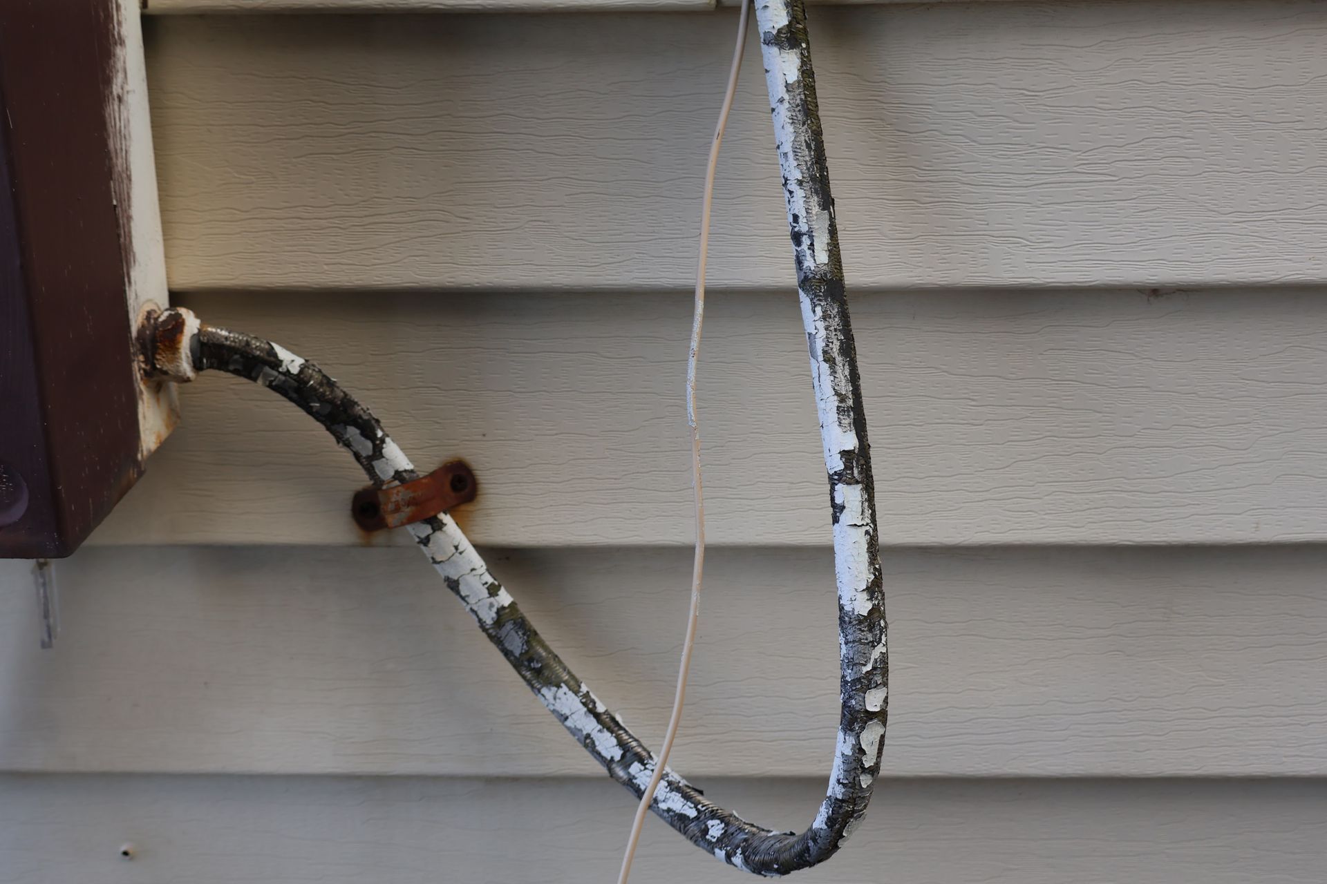 A weathered, white-painted conduit pipe attached to light-colored horizontal siding, featuring a rusted metal strap mount.