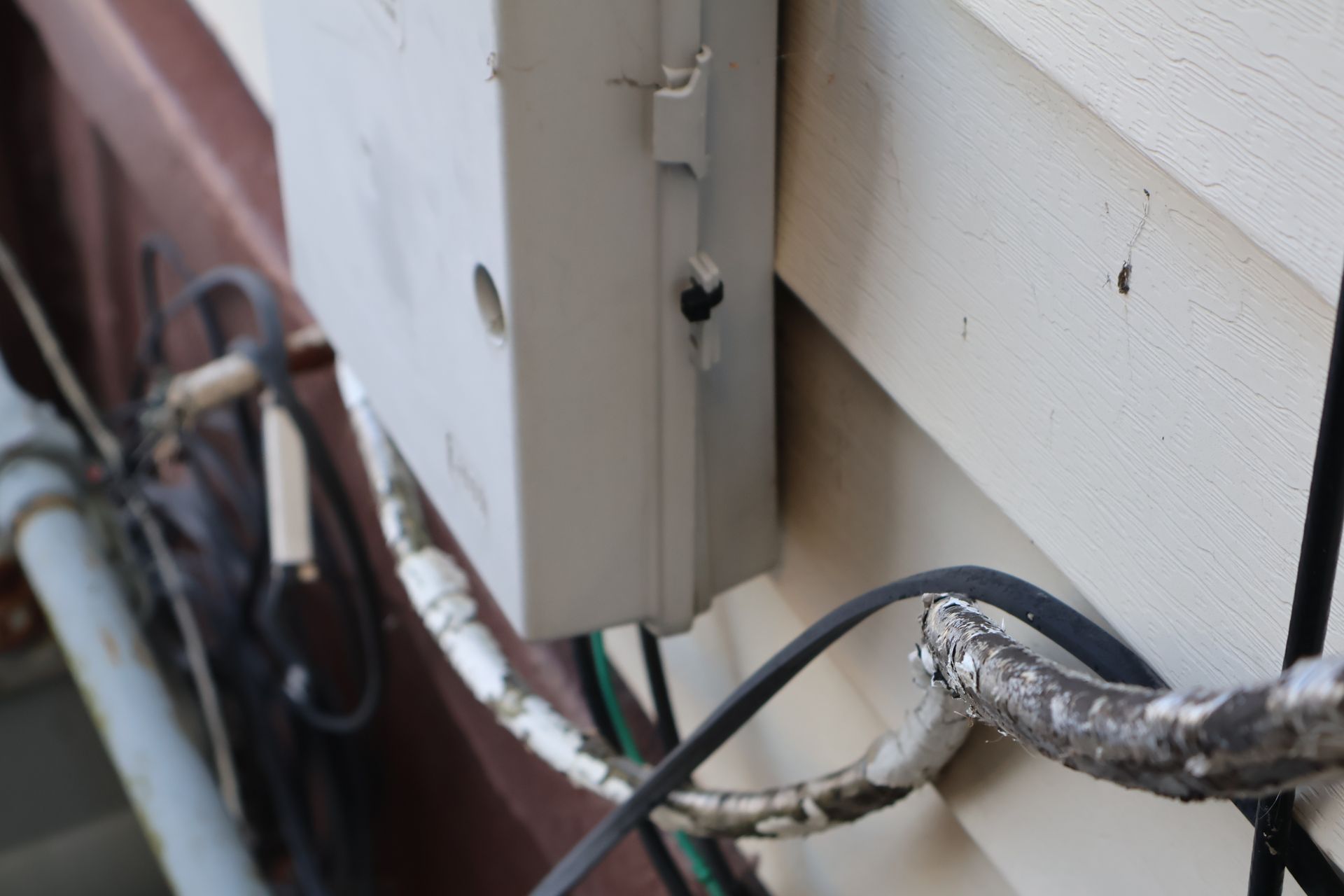 A light gray utility box mounted on beige siding with several exposed cables and wires running along the wall below.
