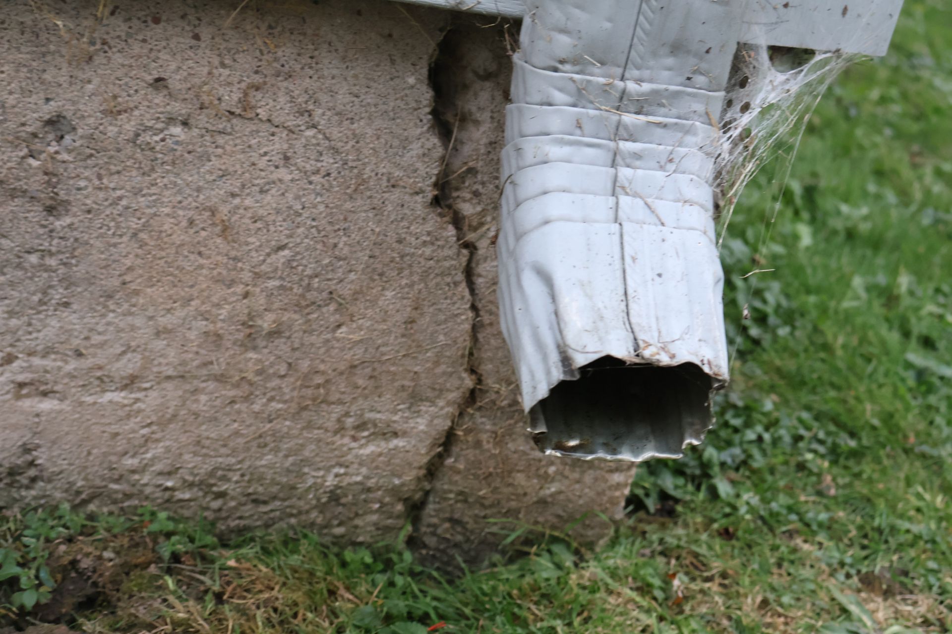 A white downspout positioned directly next to a vertical crack in a concrete house foundation near the ground.
