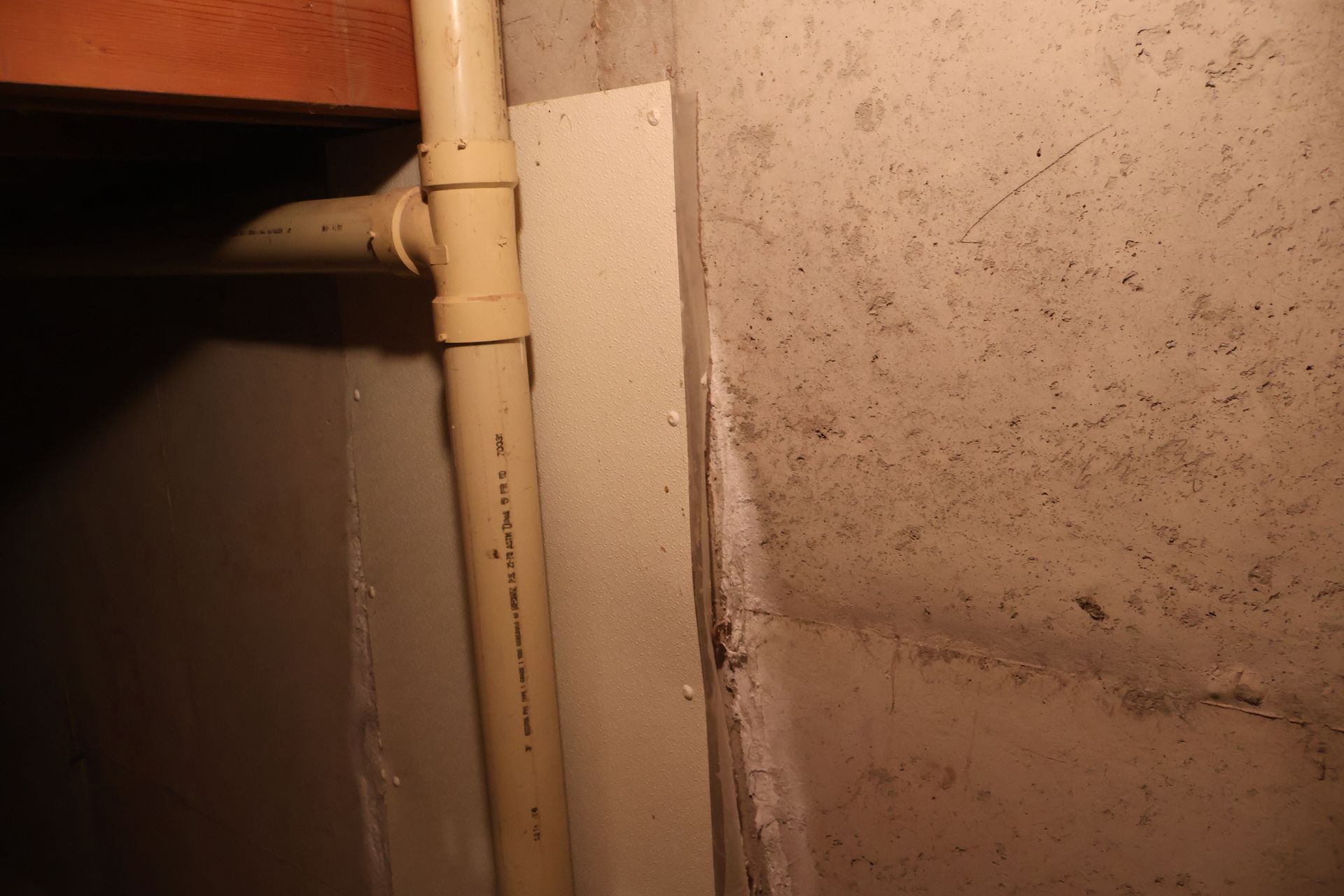 A vertical beige pipe runs in front of a flat board next to a rough, concrete wall in a dimly lit basement area.