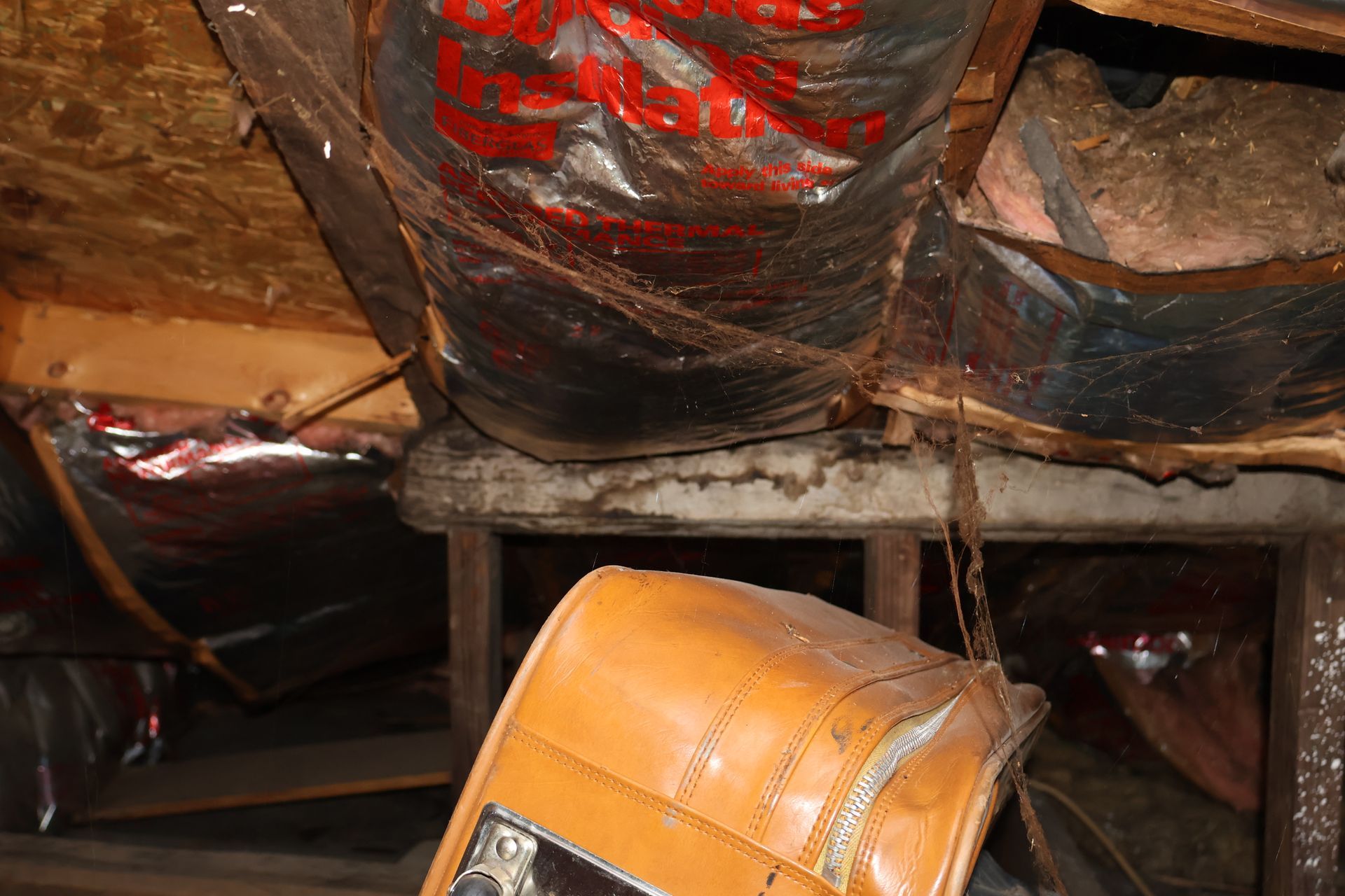 A view inside an attic showing damaged silver ductwork and insulation, with a light-brown leather suitcase in the foreground.