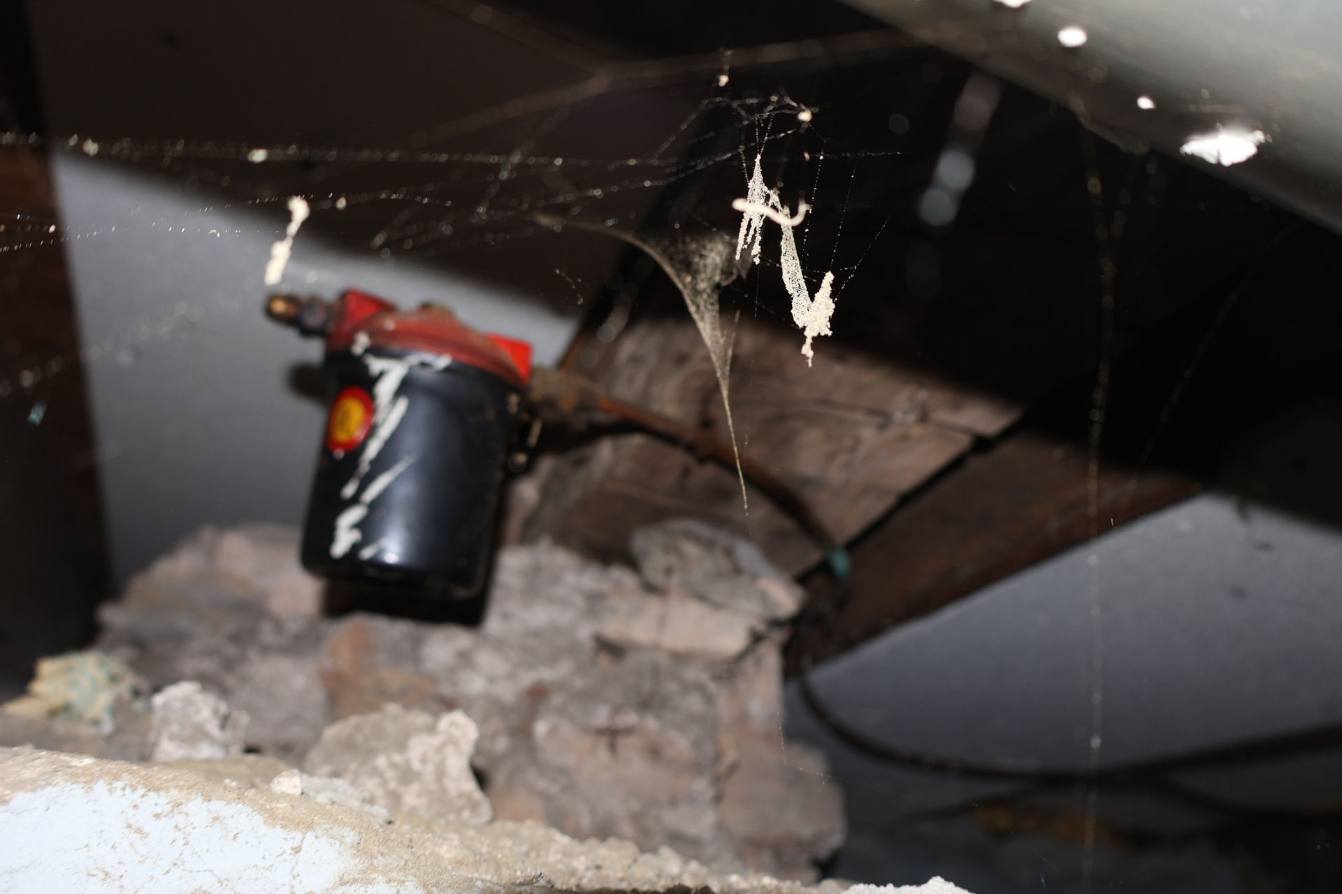 A black oil filter with a red top sits on a pile of debris beneath a wooden beam in a dimly lit, cobweb-filled space.