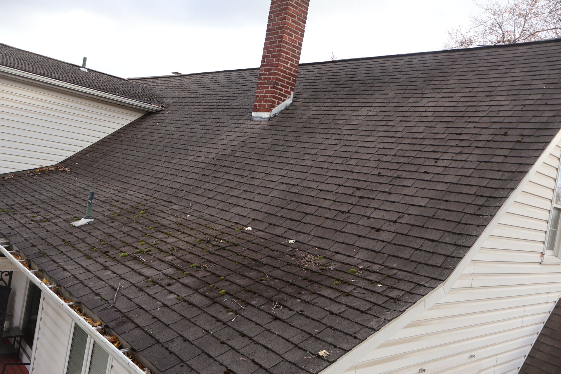 An elevated view of a dark shingled roof with a brick chimney and debris accumulation near the eaves.