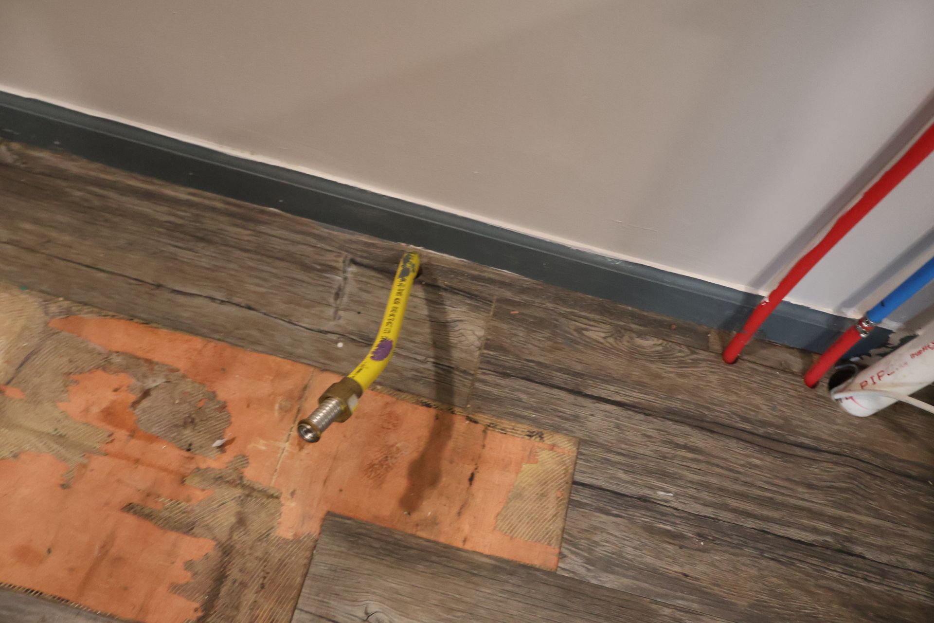 A yellow gas pipe stub protruding through a floor with wood-look planks and adjacent red and blue water supply lines.