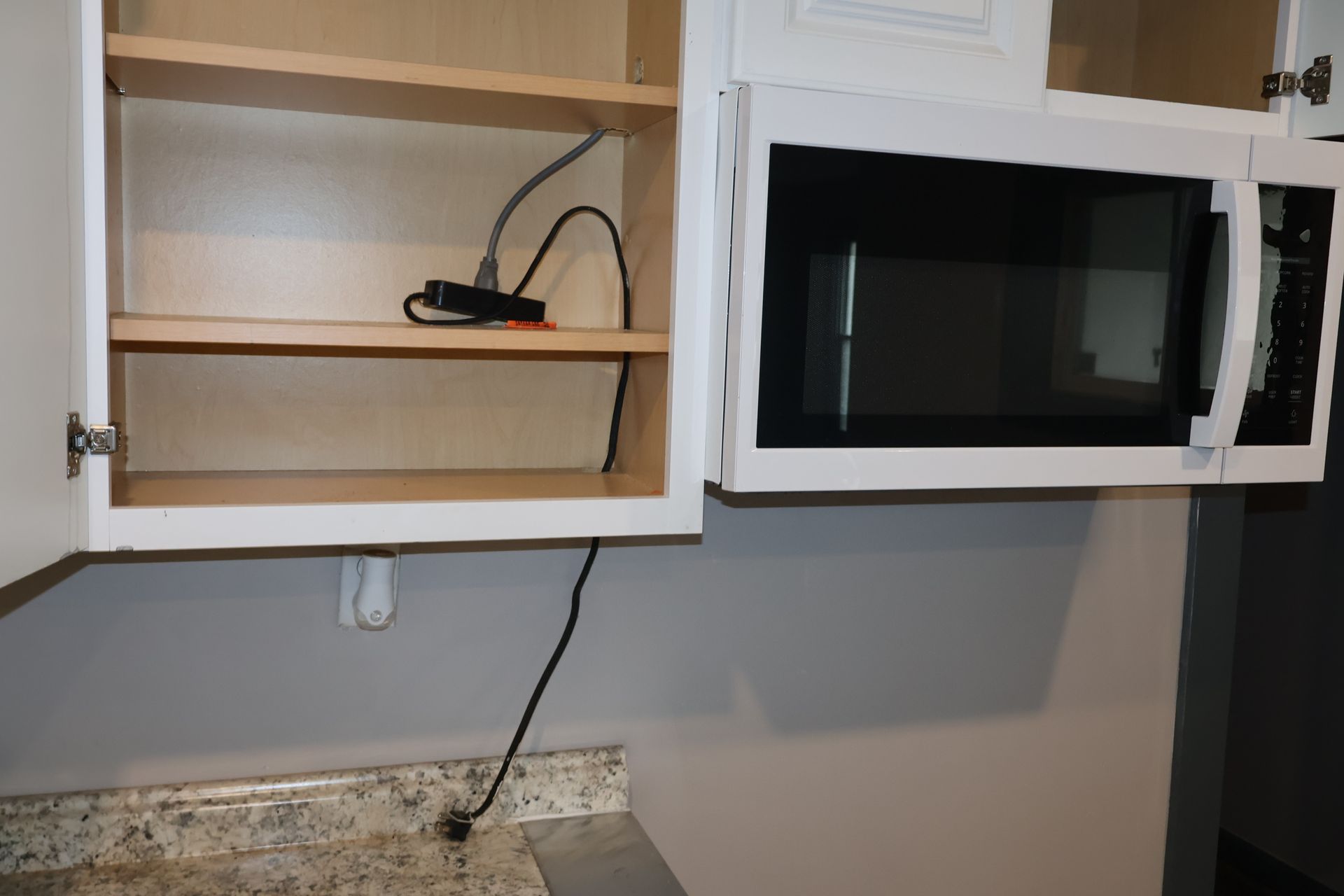 A white cabinet with shelves and a mounted microwave, with a power cord hanging down in front of the kitchen wall.