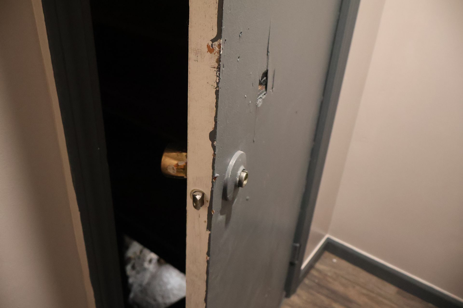 A slightly open grey door with a visible deadbolt and damaged paint on the door frame in an indoor hallway.