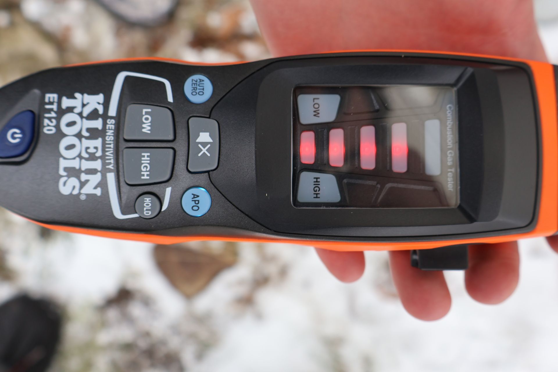 A hand holds an orange and black Klein Tools ET120 gas leak detector showing four red light bars on the display.