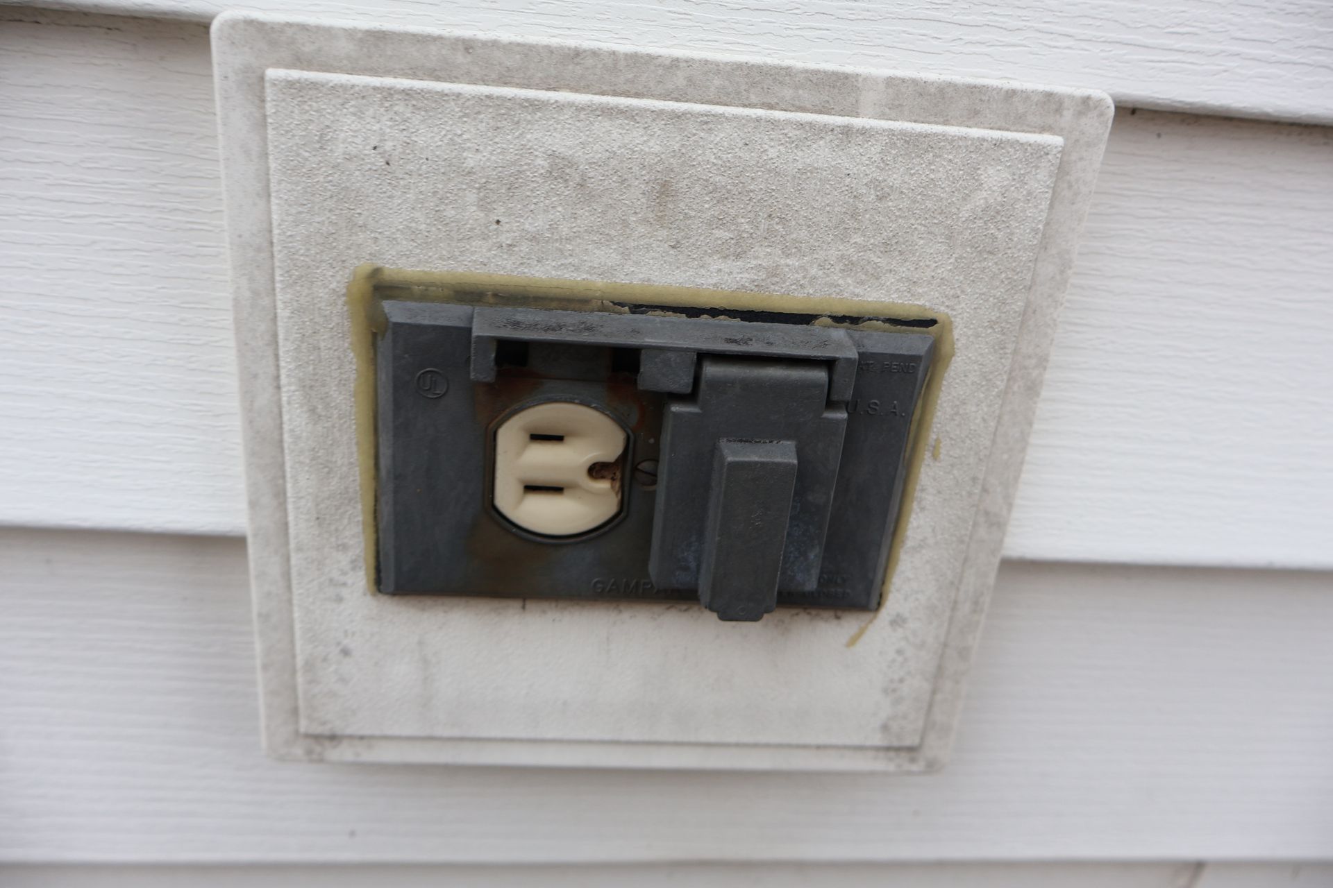 An outdoor electrical outlet with a weather-resistant gray cover mounted on white siding.