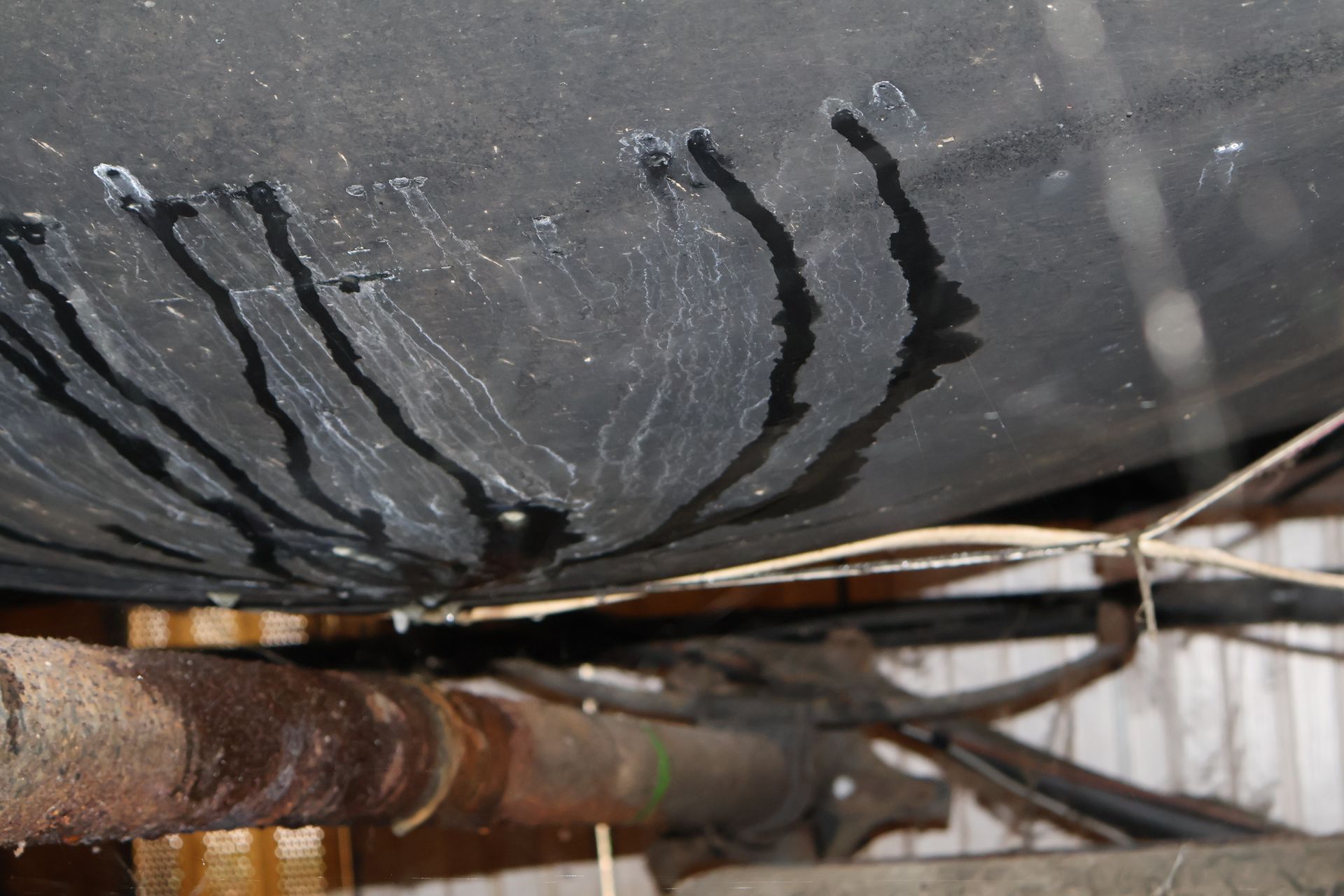 Dark, leaking liquid streaks downward on the underside of a black container, above rusted metal machinery.