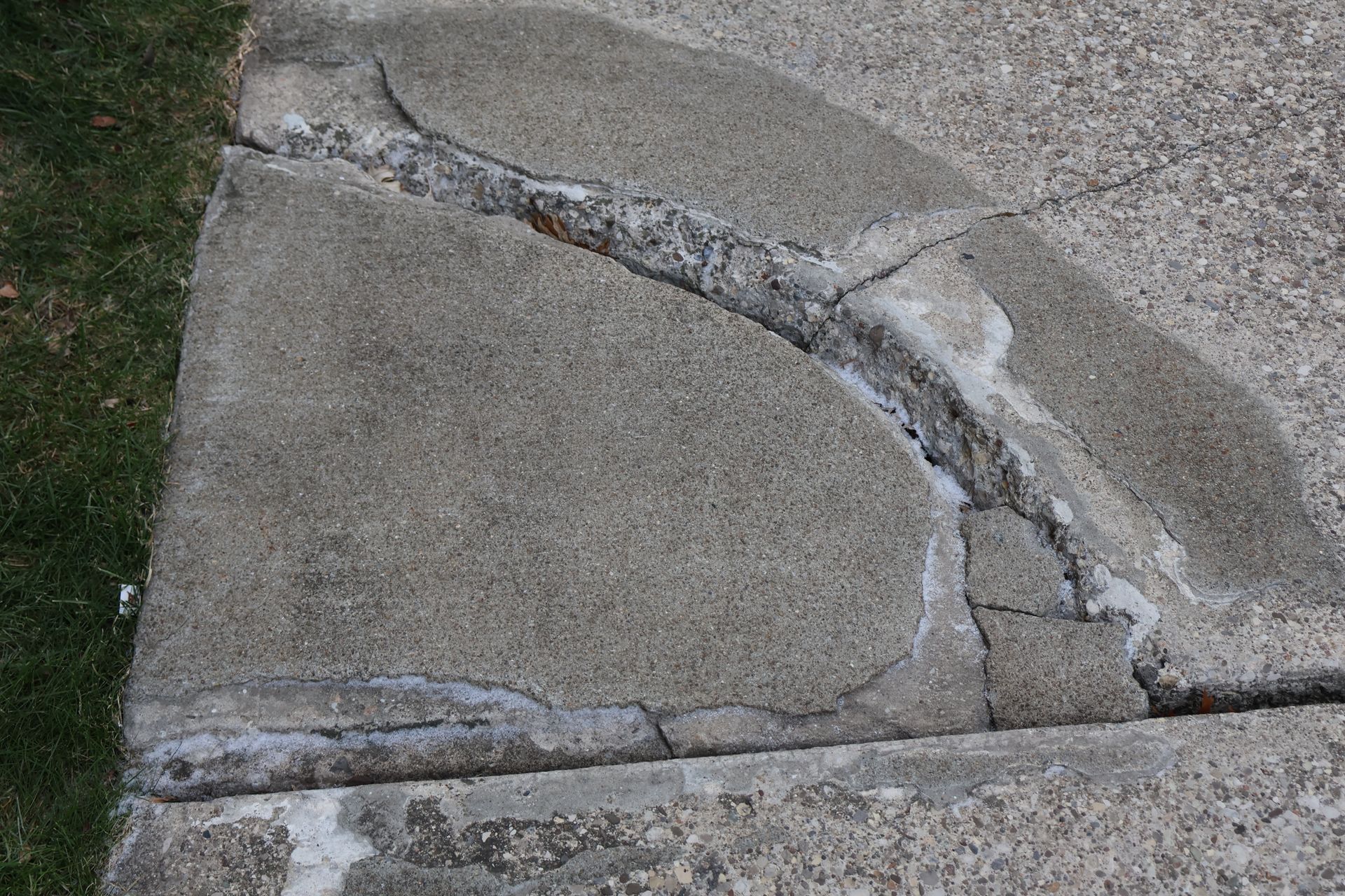 A large, jagged crack runs through a textured grey concrete sidewalk, showing signs of previous patch repairs.