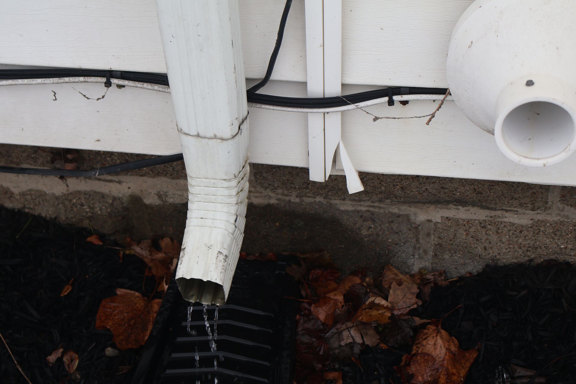 A white downspout drains water into a black grate on the ground next to a white exterior wall and a circular vent.