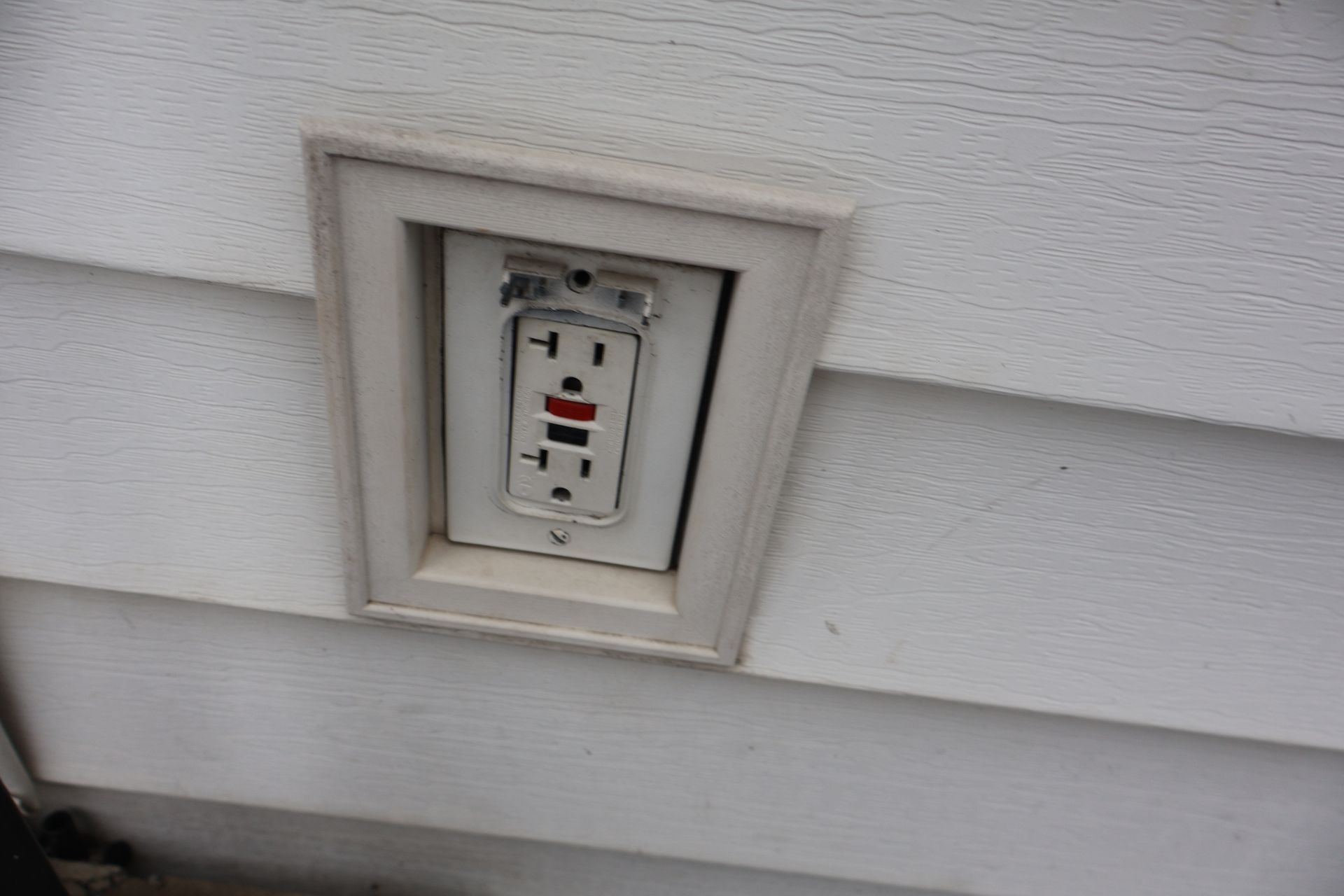An outdoor GFCI electrical outlet installed in white siding, missing its protective weather cover.
