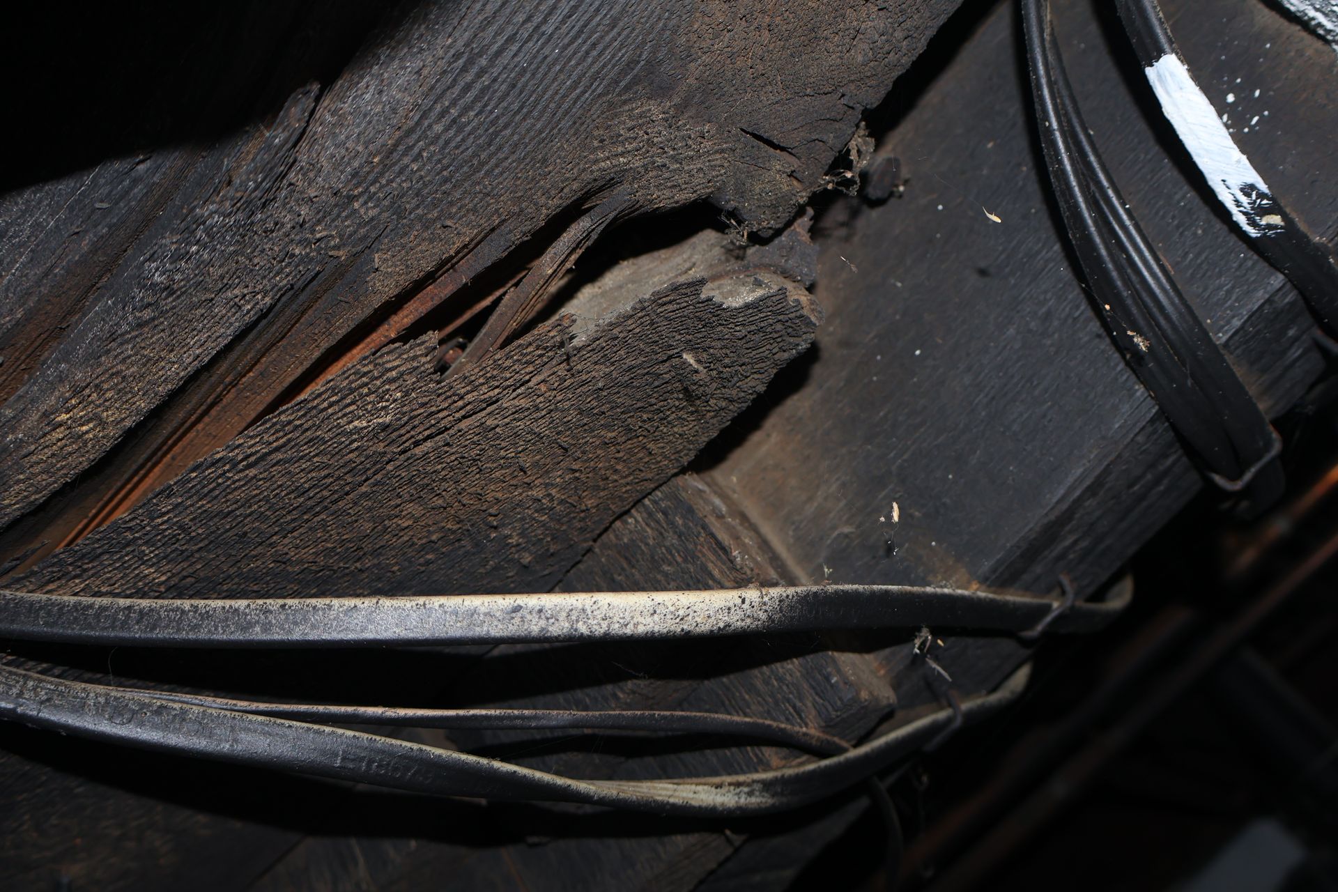 A dark, wooden structural beam with visible splintering, decay, and frayed electrical cables running across its surface.