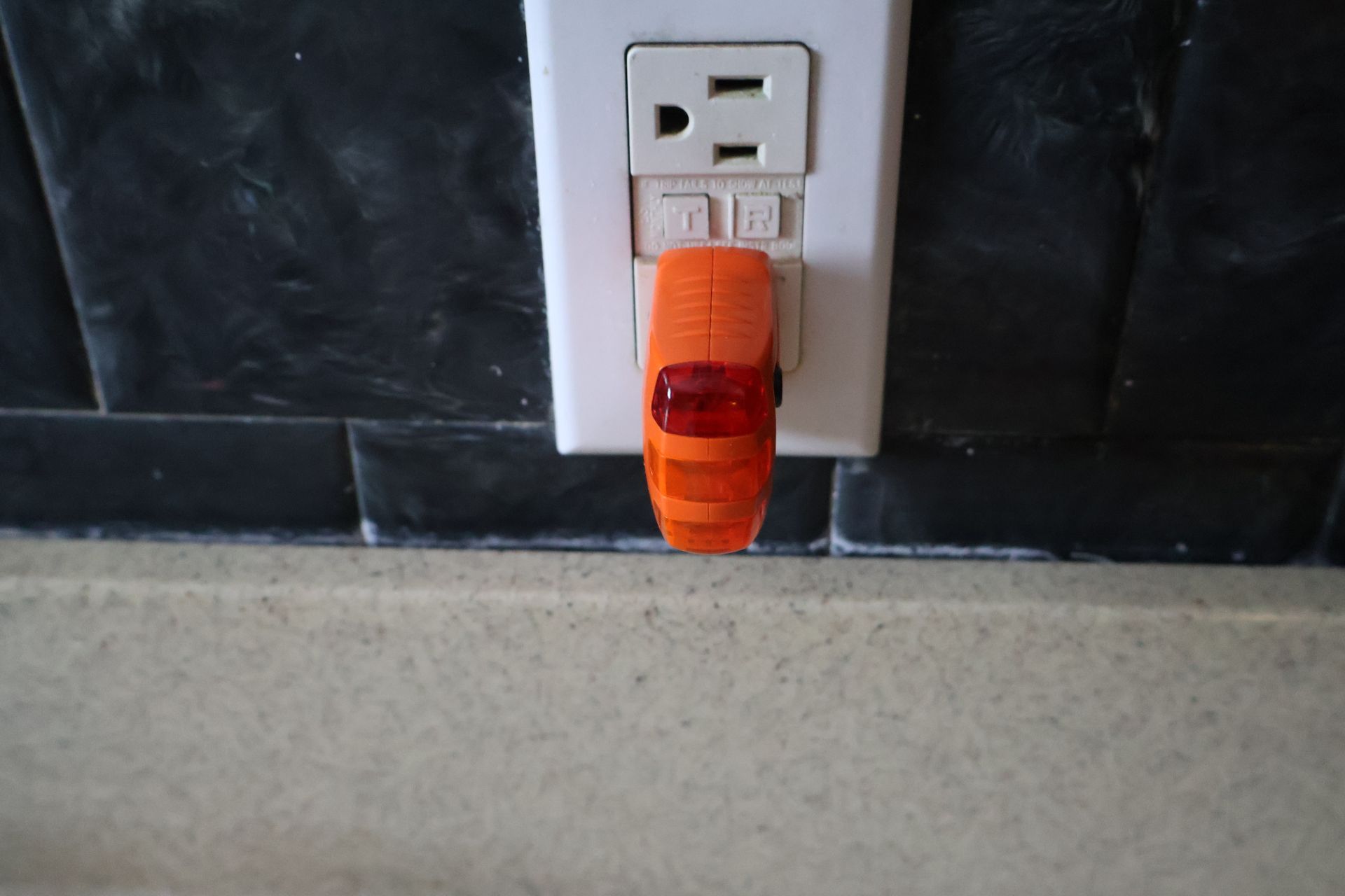 An orange plug-in nightlight is inserted into a white GFCI outlet on a dark tiled kitchen backsplash above a countertop.