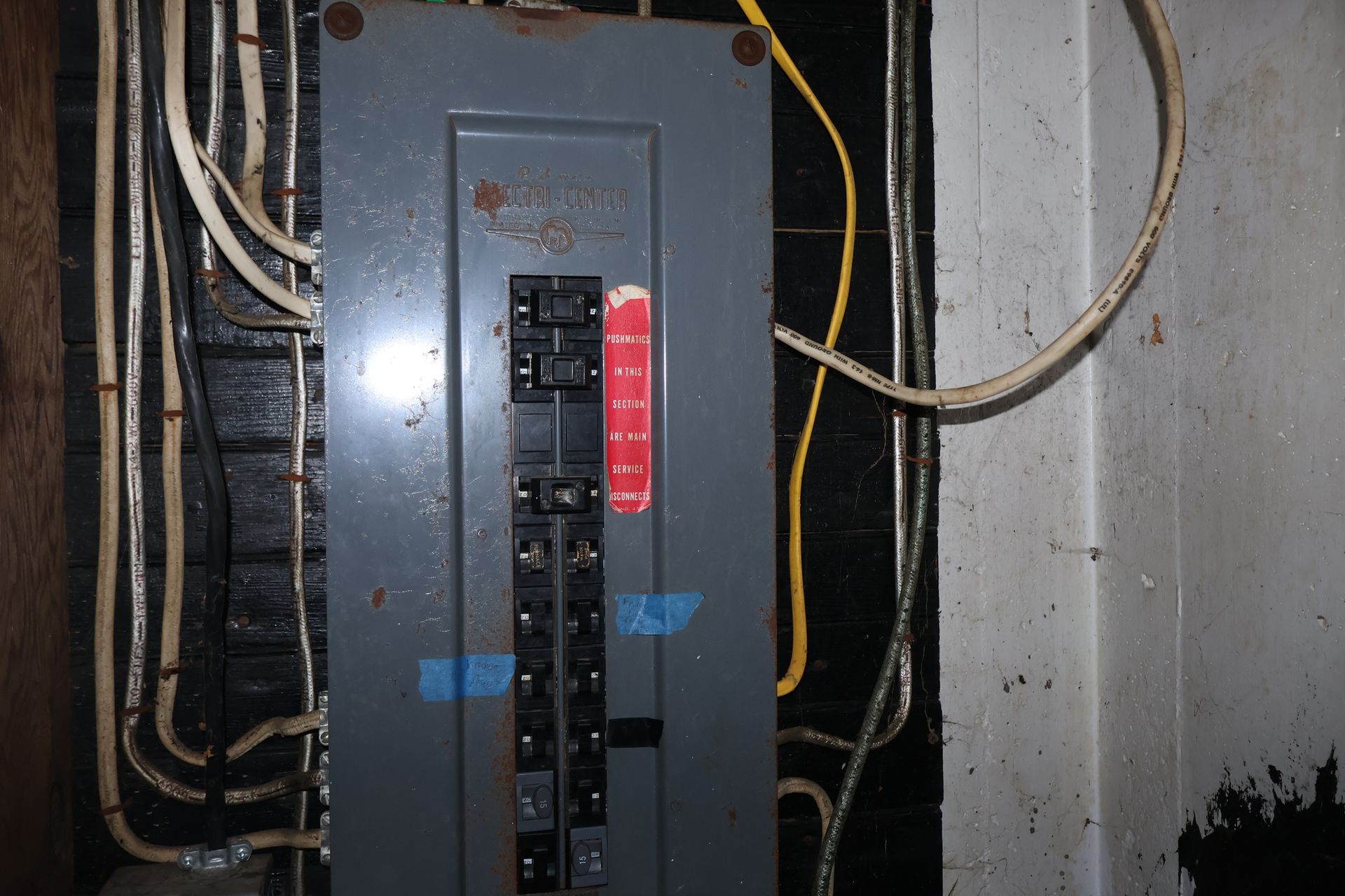 Electrical breaker panel mounted on a dark wooden wall with multiple white and yellow cables connected to the top.