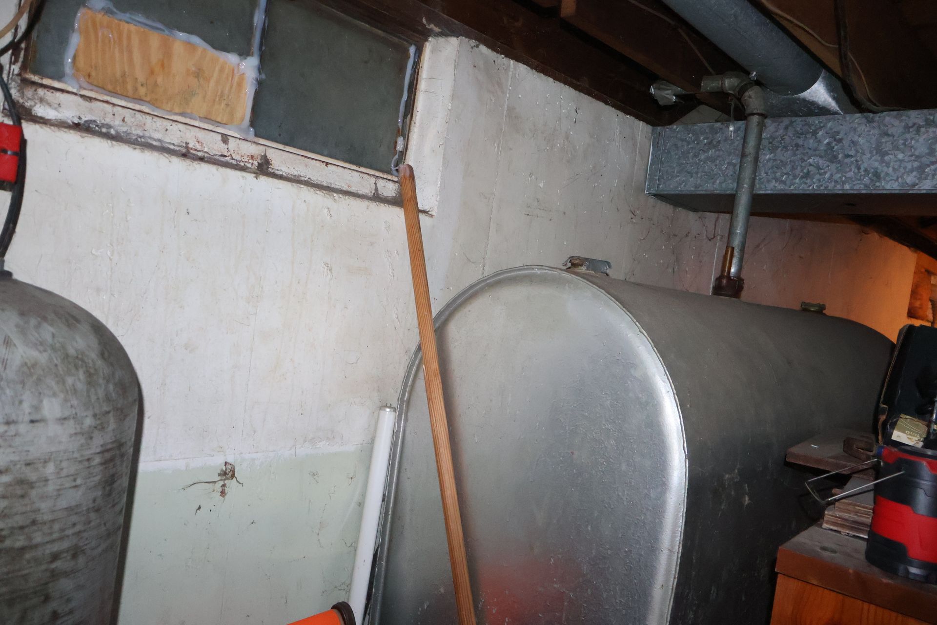 A basement wall with an oil tank, a window partially covered with plywood, and a vertical metal rod leaning against the wall.