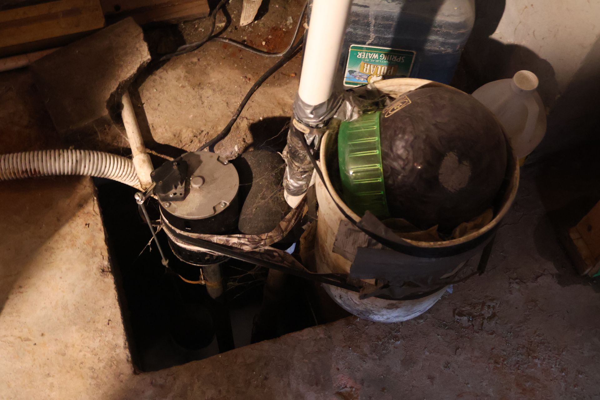 A sump pump setup in a concrete basin, featuring a pump, PVC discharge pipe, and a plastic bucket with debris.