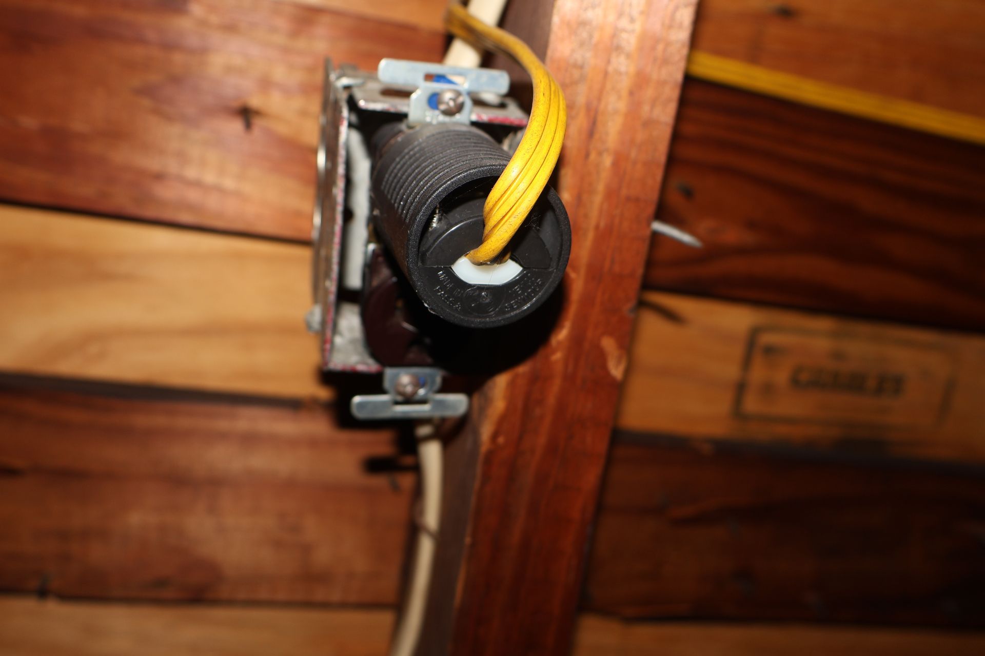 An electrical box mounted to a wooden beam with an exposed black plug and yellow extension cord wires.