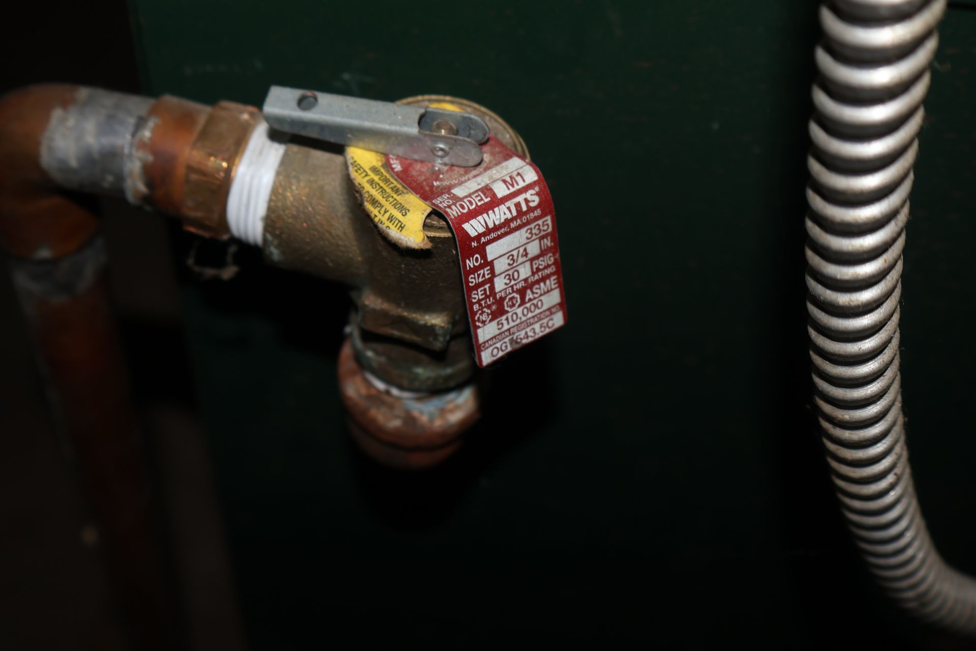 A brass Watts pressure relief valve with a red identification tag attached to piping on a dark green boiler unit.