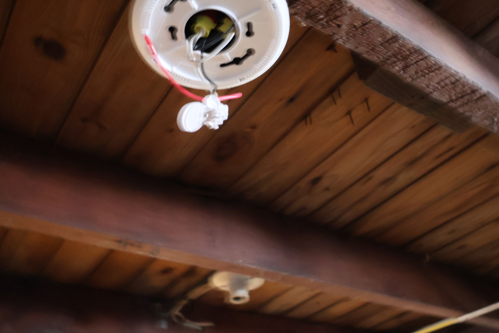 A smoke detector base plate mounted to a wooden ceiling, with loose wiring and an electrical connector hanging down.