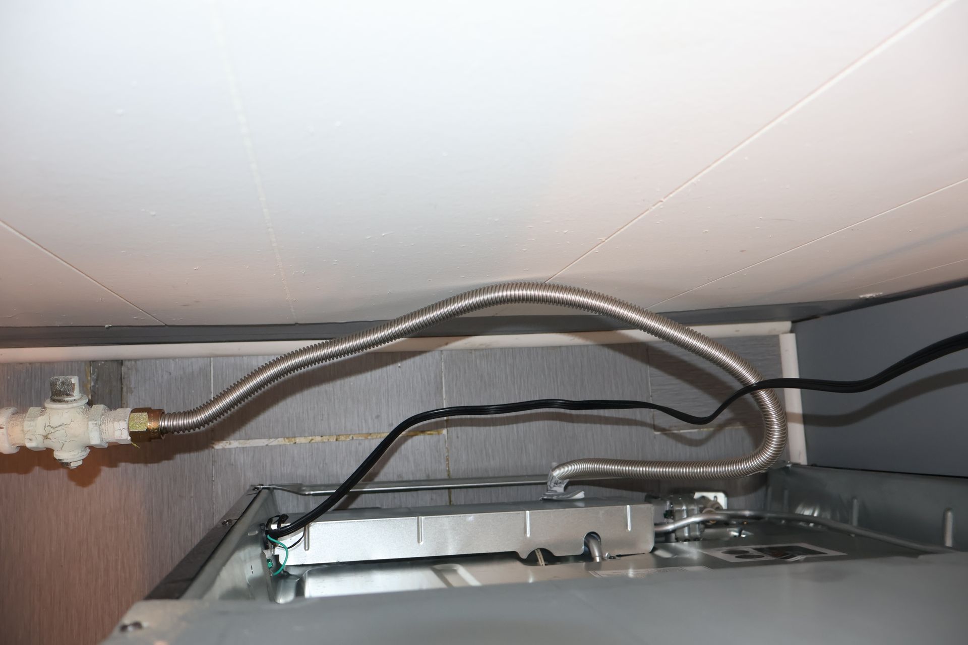 A braided metal water supply line and black electrical cord connected to a metallic appliance under a ceiling.