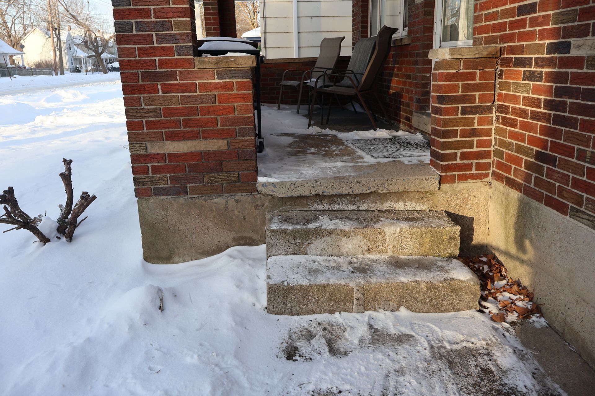 Two concrete steps lead up to a snow-covered porch with two chairs, set against a red brick house in winter.