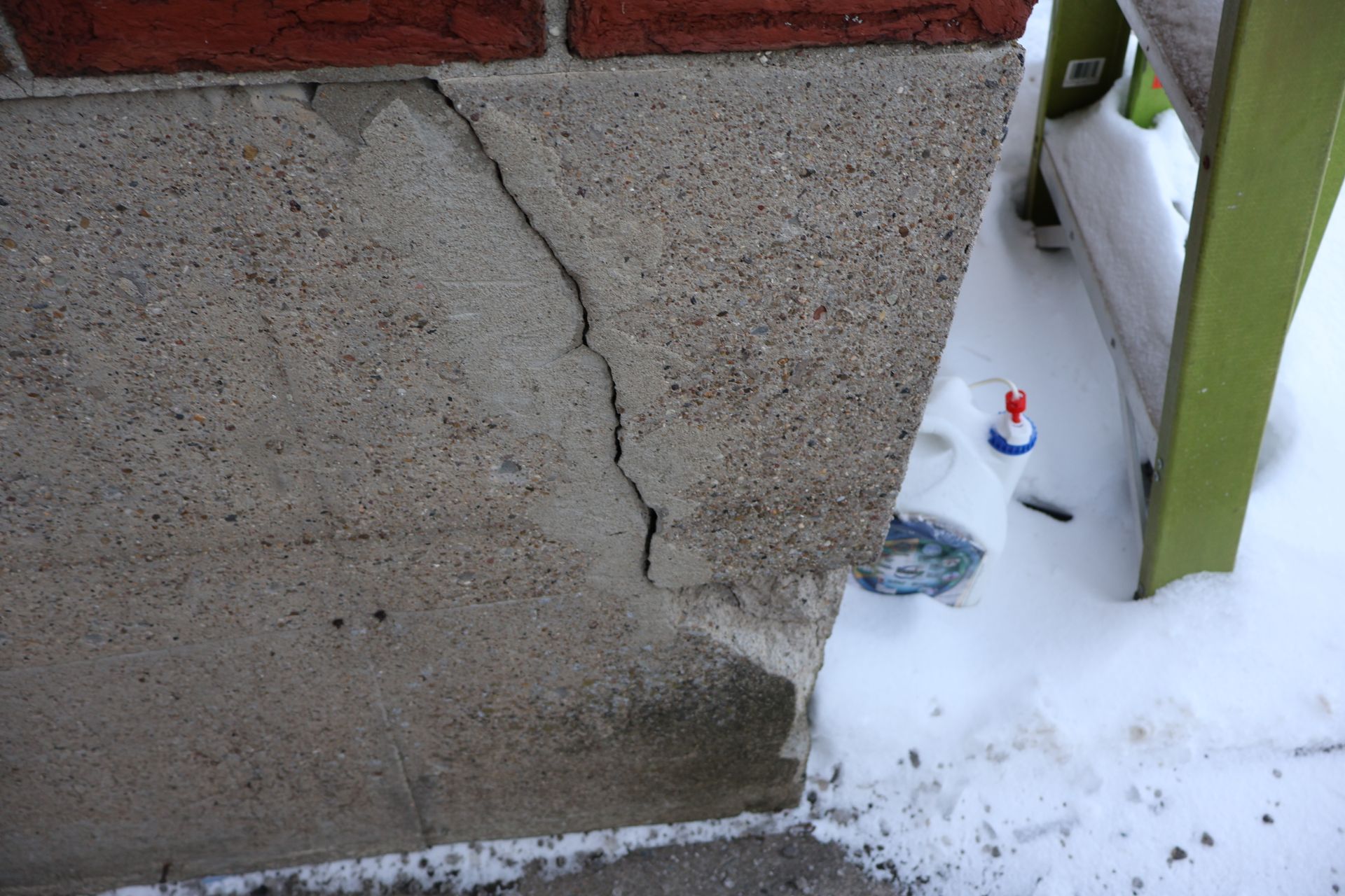 A diagonal crack runs through a gray concrete foundation wall beneath a red brick exterior, with snow on the ground.