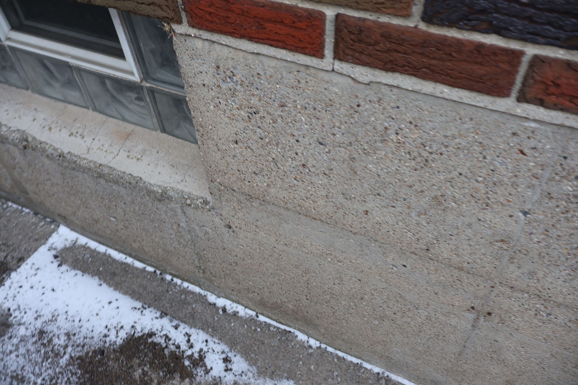 A close-up view of a concrete foundation wall with a glass block window, positioned below a brick exterior wall.