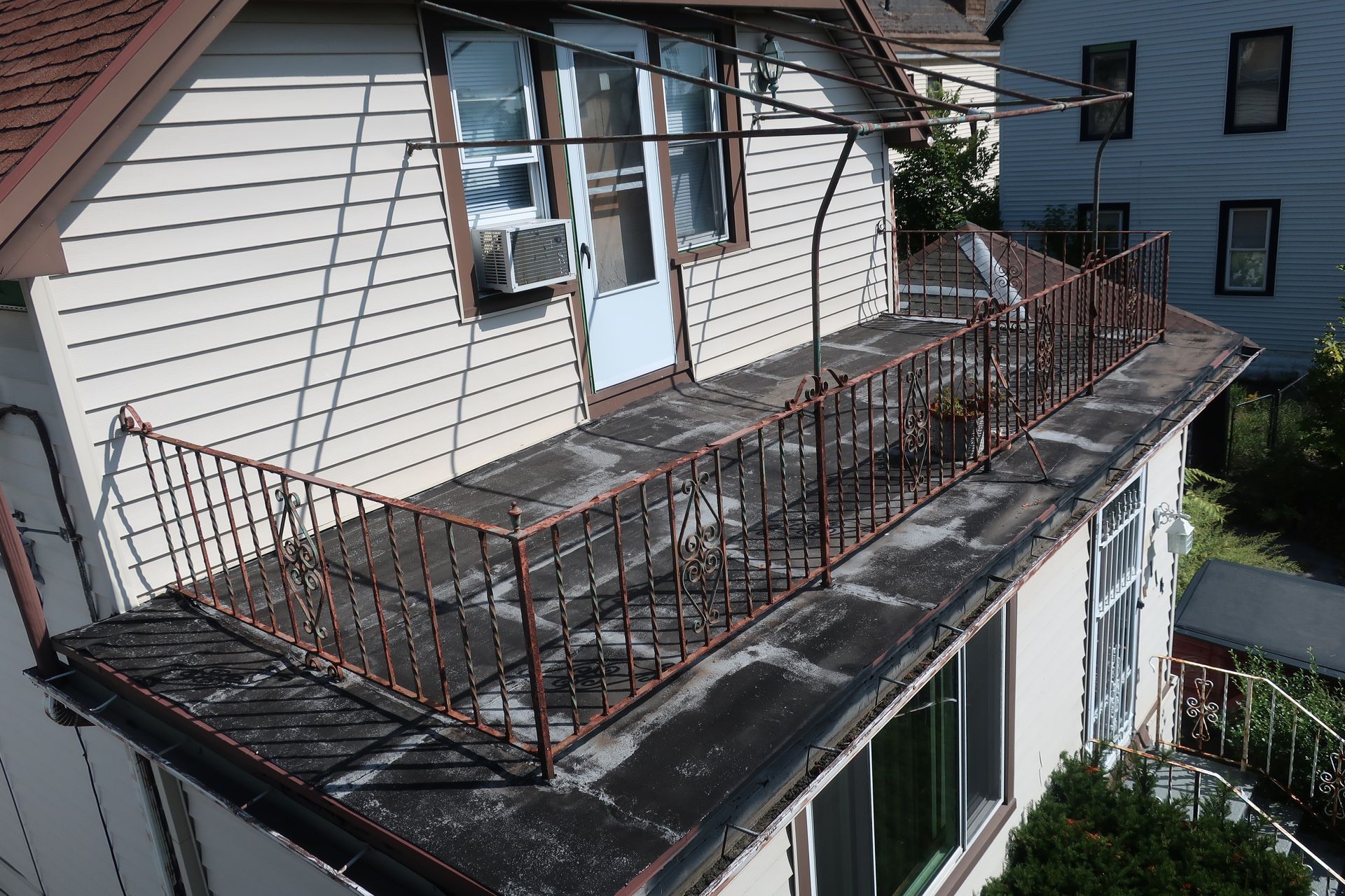 A white house features an upper-level flat rooftop deck enclosed by a rusted metal railing and a metal canopy frame.