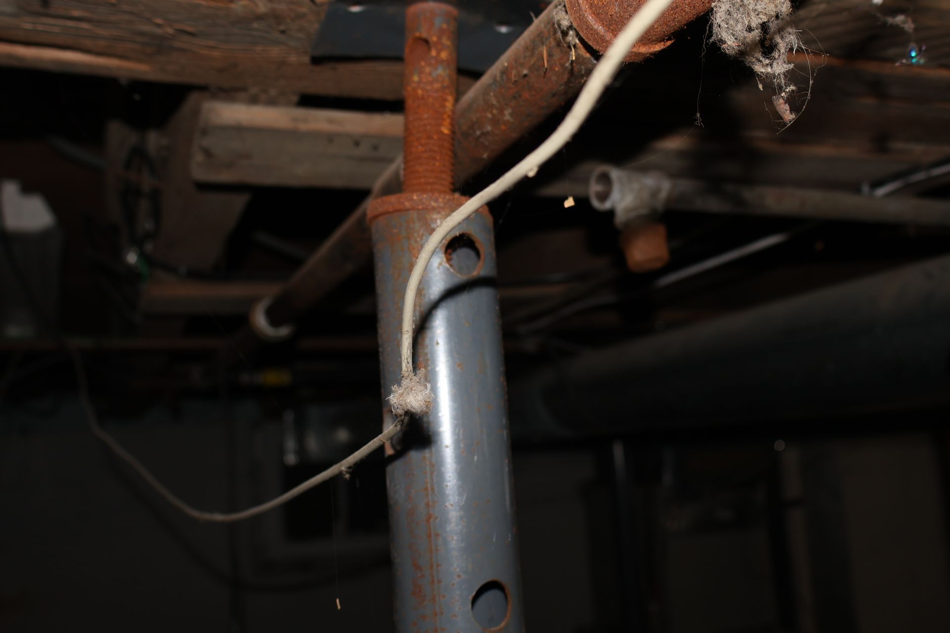 A rusty metal adjustable support column in a dimly lit basement with a thin white wire draped across it.