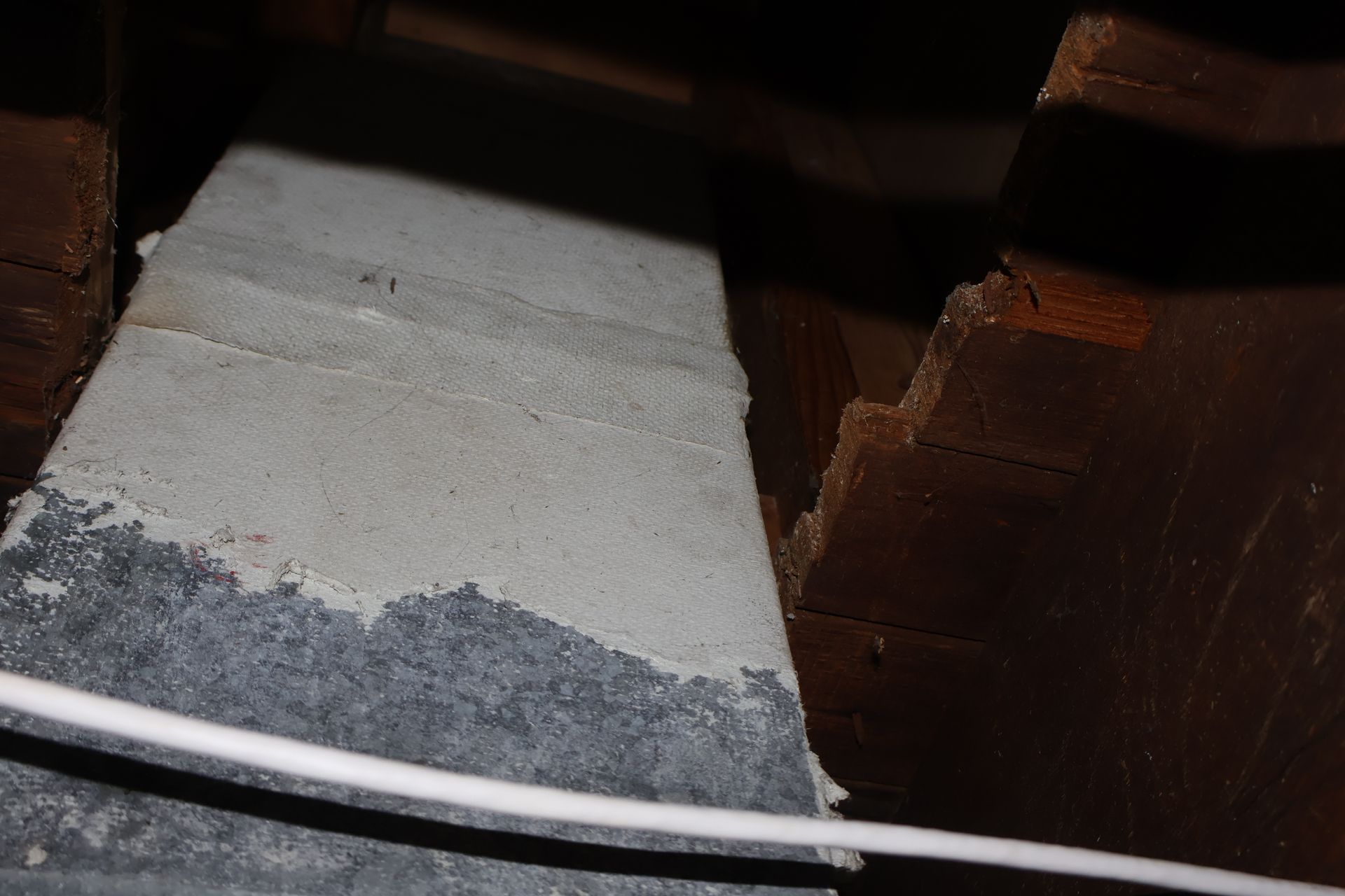 A gray metal rectangular duct runs through a dark crawlspace beside rough, unfinished wooden beams.