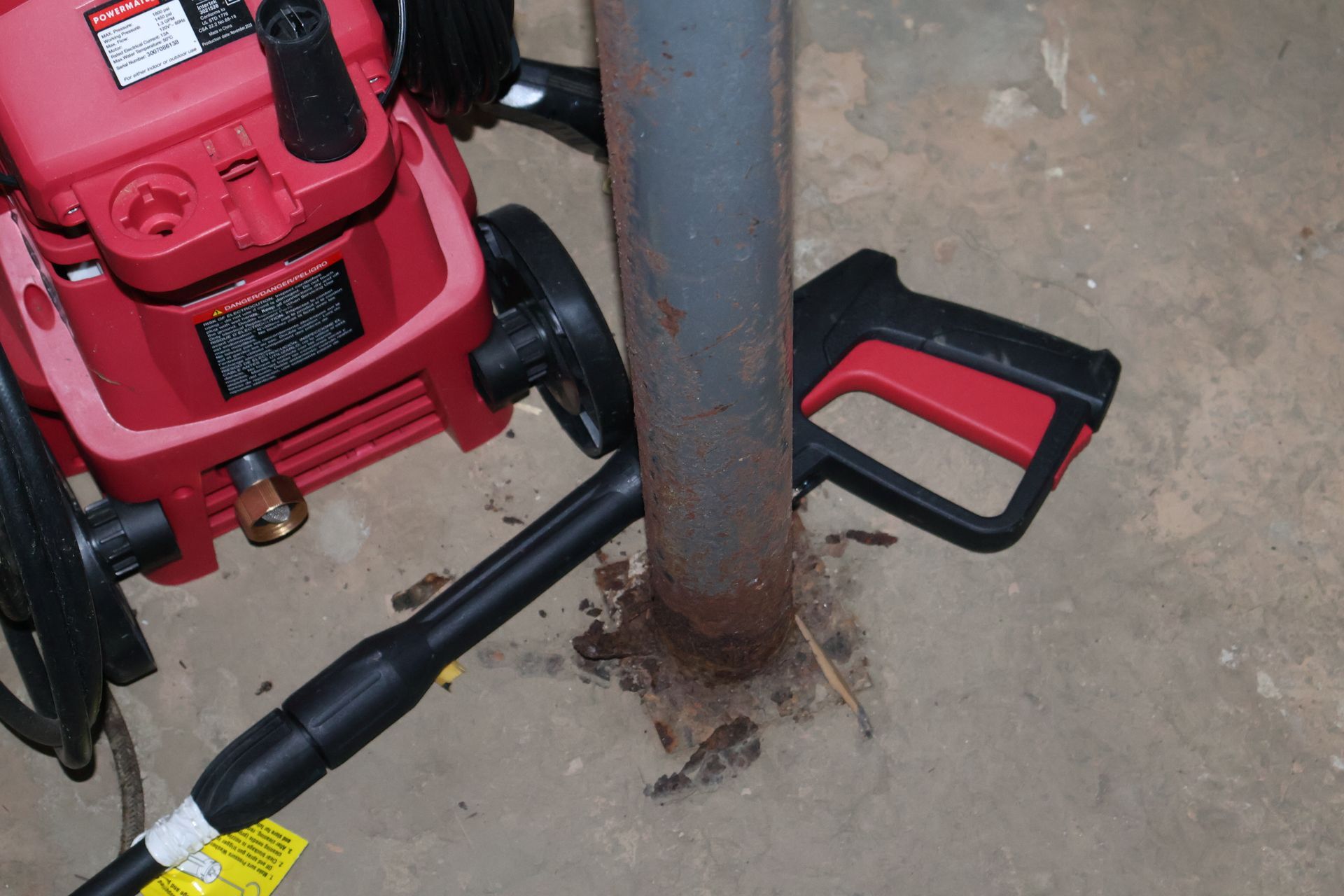 A red pressure washer rests on a concrete floor next to a rusted metal pole with its spray gun lying on the ground.