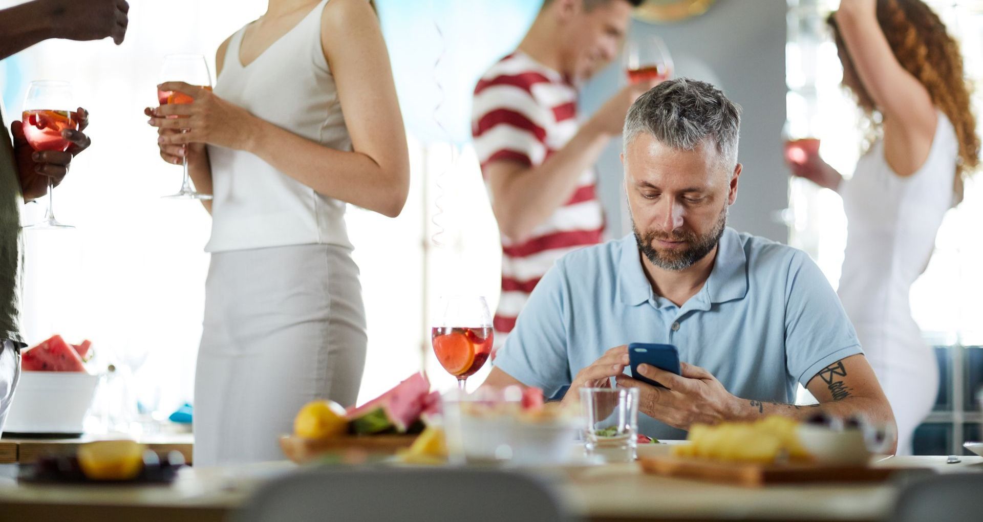 Man at party looks at his phone while others mingle, holding drinks.