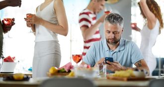 Man at party looks at his phone while others mingle, holding drinks.
