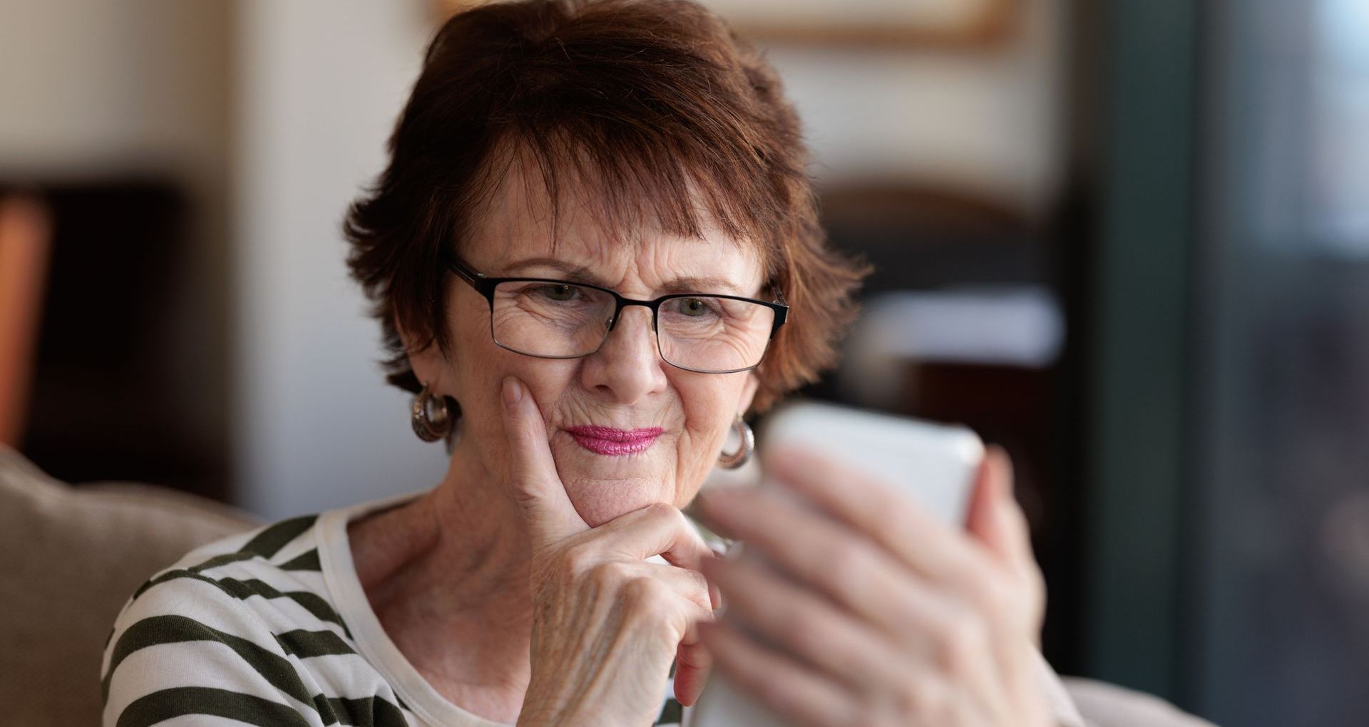 Woman in glasses holding smartphone, looking at the screen with a thoughtful expression. Indoors.