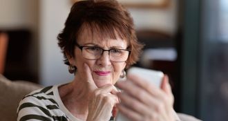 Woman in glasses holding smartphone, looking at the screen with a thoughtful expression. Indoors.