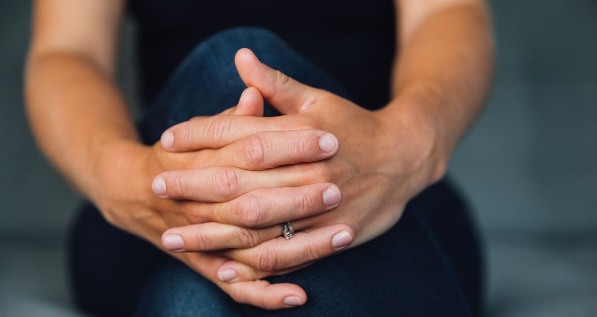 Person’s hands clasped in lap; wearing a ring. Against a blurry background.