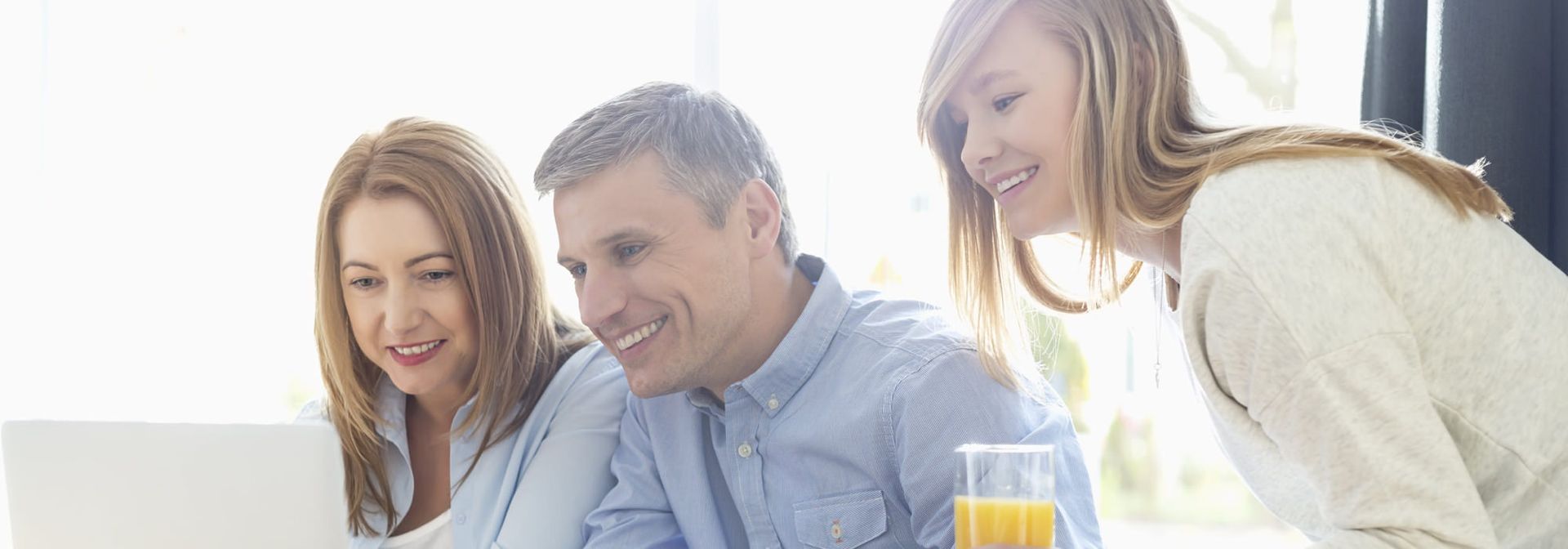 Three people smile while looking at a laptop screen. A glass of orange juice sits to the right.