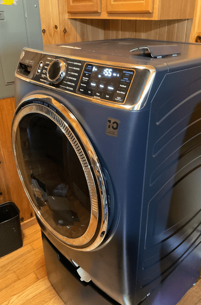 Blue washing machine in a wood-paneled room. It has a chrome door and control panel.