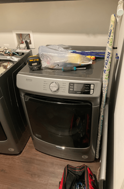 A gray dryer in a laundry room, with tools on top. The door is closed, and there's a red tool bag below.