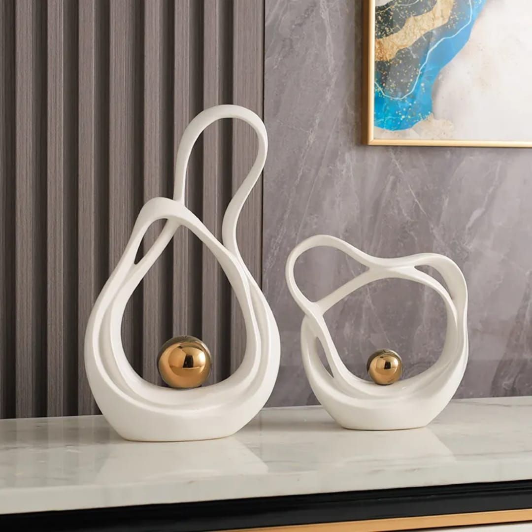Two white abstract loop sculptures with gold accents on a marble shelf against a gray textured wall.