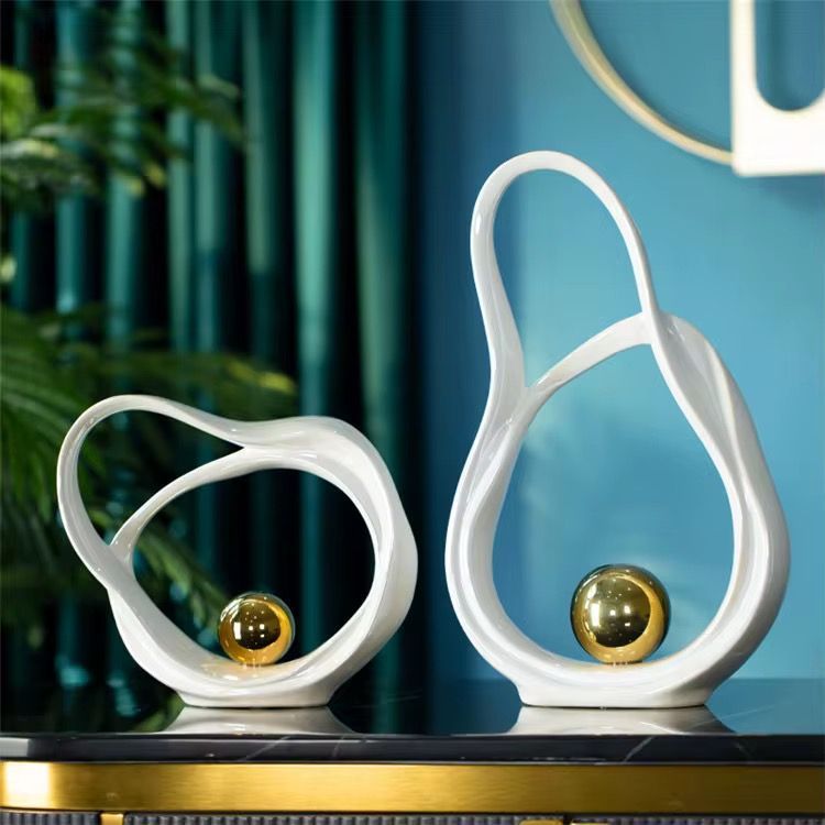 Two white looped sculptures with gold spheres on a shelf against a teal wall.