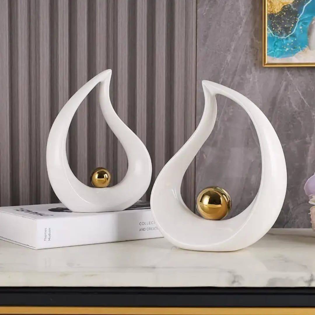 Abstract white flame-shaped sculpture with a gold sphere on a marble surface, beside flowers
