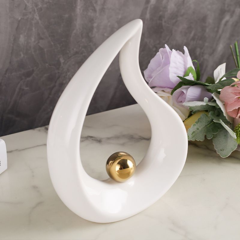 Abstract white flame-shaped sculpture with a gold sphere on a marble surface, beside flowers