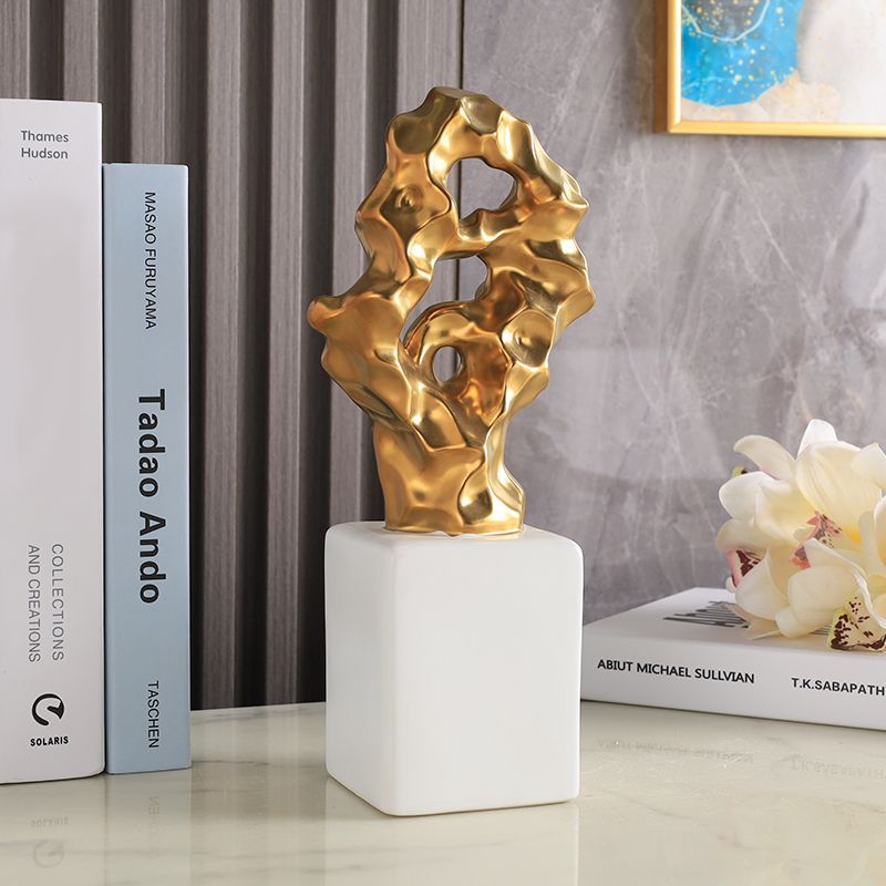 Gold abstract sculpture on a white pedestal beside books and white flowers on a shelf