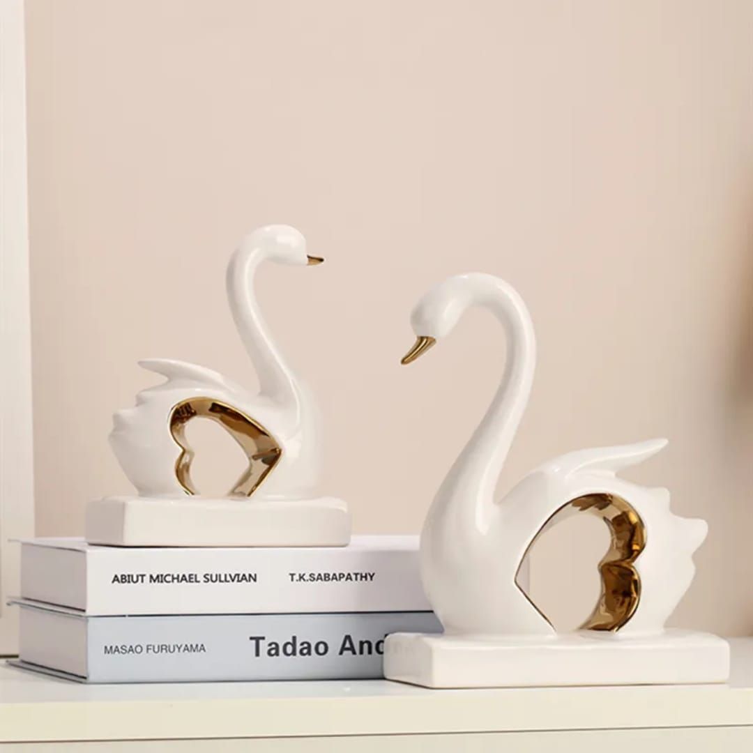 Two white swan-shaped ceramic ornaments with gold accents on a white shelf, in small and large sizes