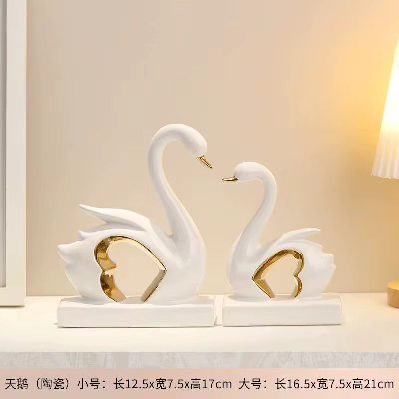 Two white swan-shaped ceramic ornaments with gold accents on a white shelf, in small and large sizes