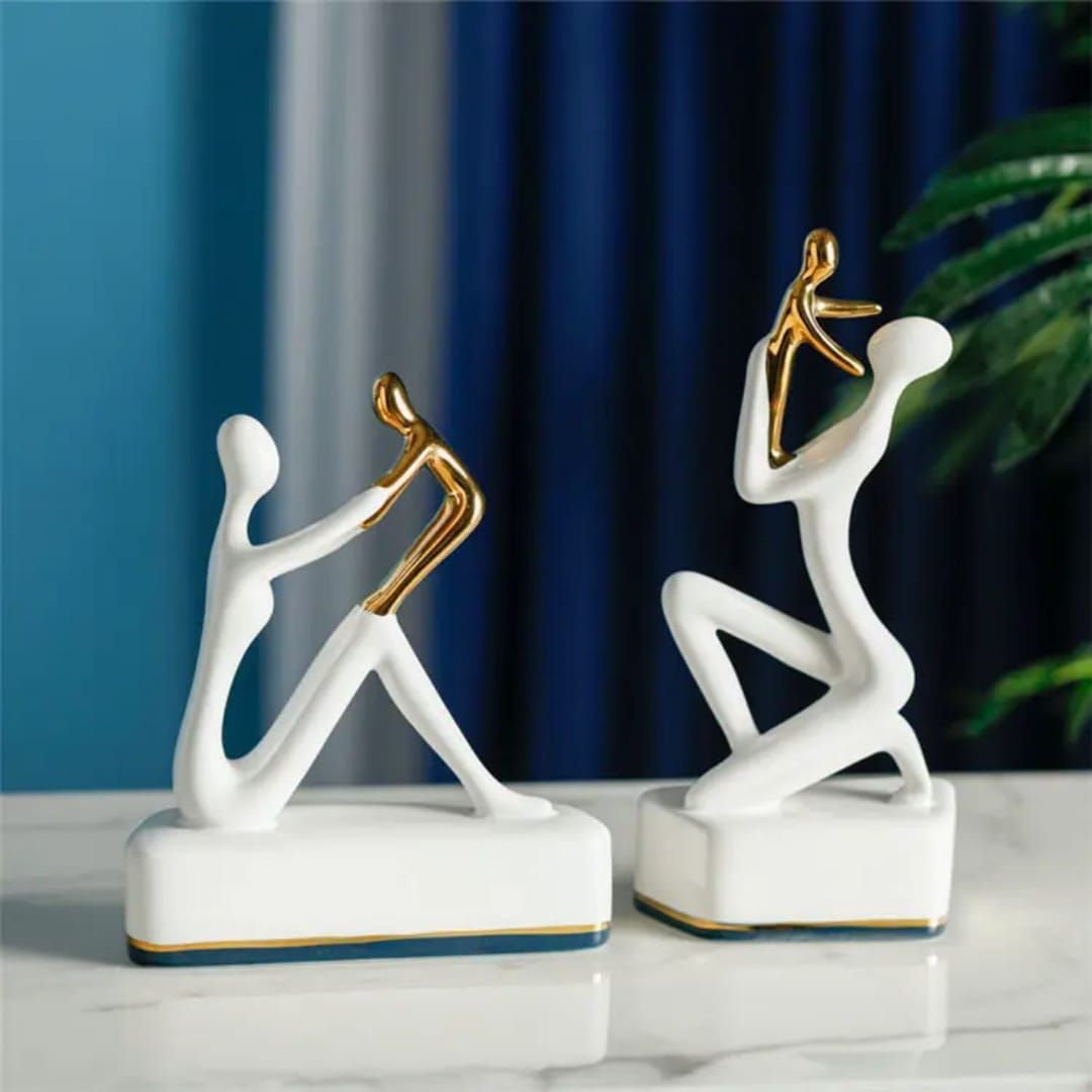 Two abstract white-and-gold figurines on a glossy table against blue curtains