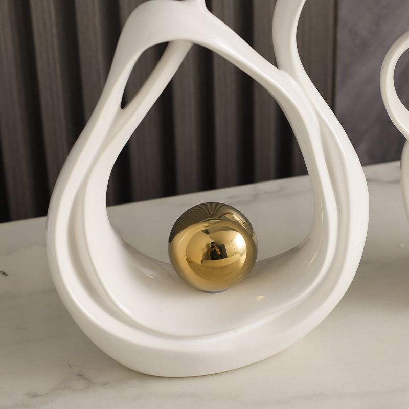 White abstract sculpture holding a gold sphere on a marble surface against a dark backdrop